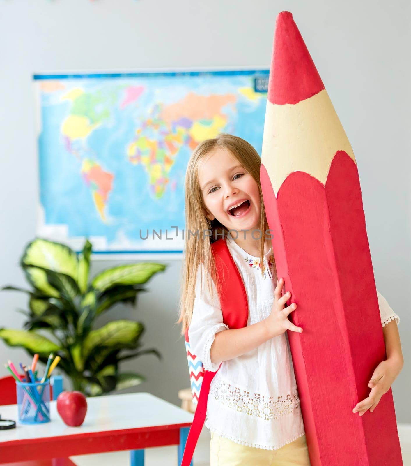 Little smiling blond girl holding huge red pencil in the school classroom by tan4ikk1