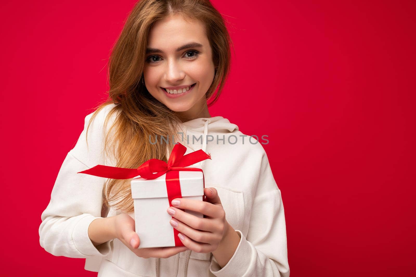 Photo shot of attractive happy smiling cute blonde young female person isolated over red background wall wearing white hoodie holding white gift box with red ribbon and looking at camera by TRMK