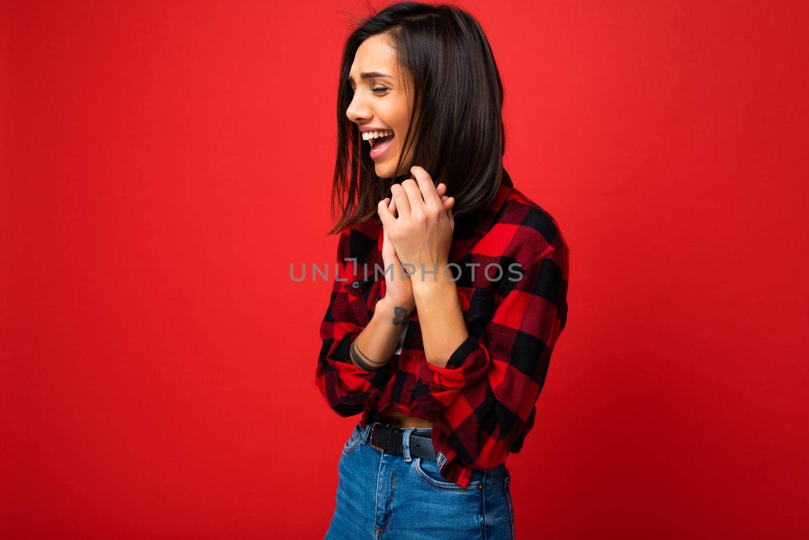 Photo of young beautiful happy smiling brunette woman wearing trendy white t-shirt and red check shirt . Sexy carefree female person posing isolated near red wall in studio with free space. Positive model with natural makeup by TRMK