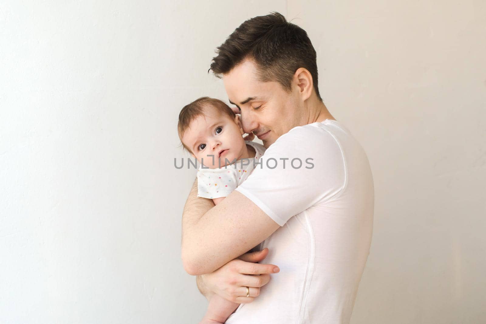 Side view of handsome young man embracing sweet baby and keeping eyes closed while standing on white background
