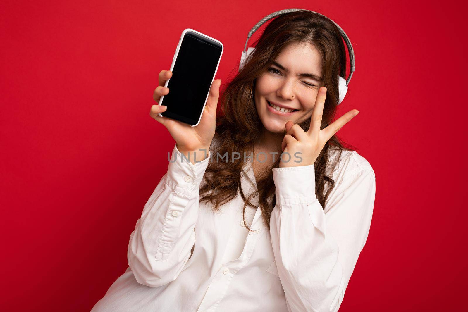 Sexy attractive positive young brunette curly woman wearing white shirt isolated on red background wall holding and showing mobile phone with empty display wearing white bluetooth headphones listening to music by TRMK