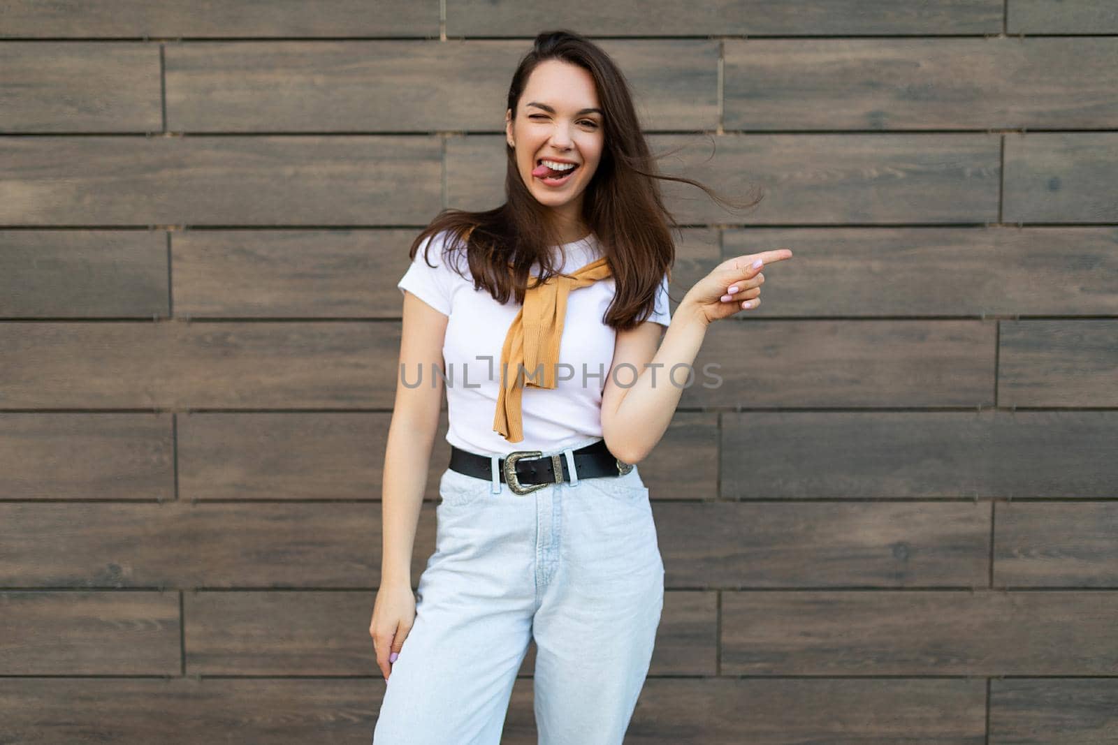 Young smiling happy winsome cute nice brunette woman wearing casual clothes standing in the street near the wall and pointing at free space. Lifestyle concept.