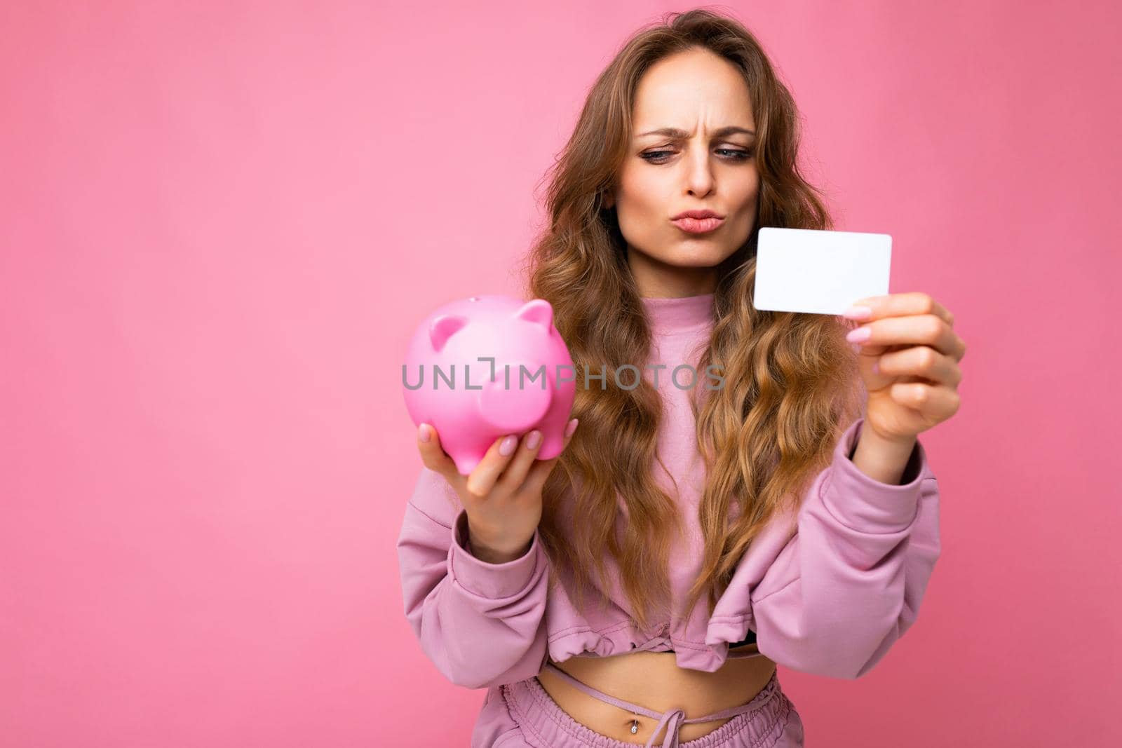 Portrait photo of doubtful asking thoughtful young beautiful attractive woman with wavy long blonde hair with sincere emotions wearing stylish pink clothes isolated over pink background with free space, holds pink piggy box and credit card. Money concept.