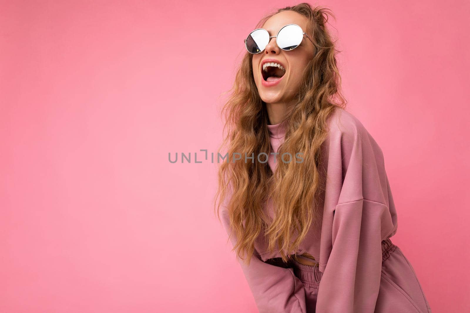 Shot of beautiful happy young dark blonde curly woman isolated over pink background wall wearing casual pink sport clothes and stylish sunglasses looking at camera and shouting. copy space