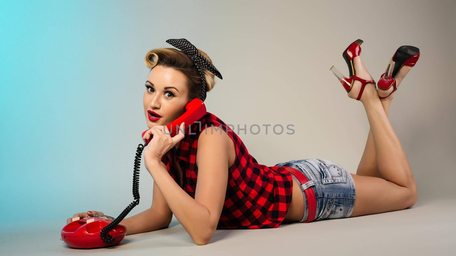Surprised attractive girl in a plaid shirt and denim shorts talking on the phone lying on a gray background. Pin up concept
