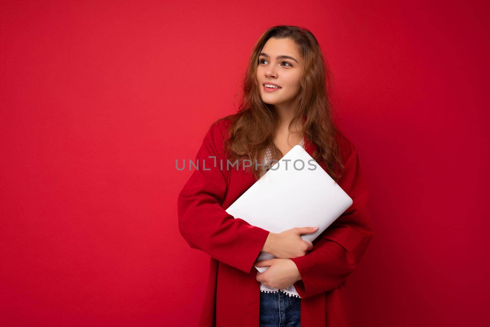 Charming mysterious young curly lady holding netbook wearing red cardigan and white blouse looking to the side isolated over red background. Copy space
