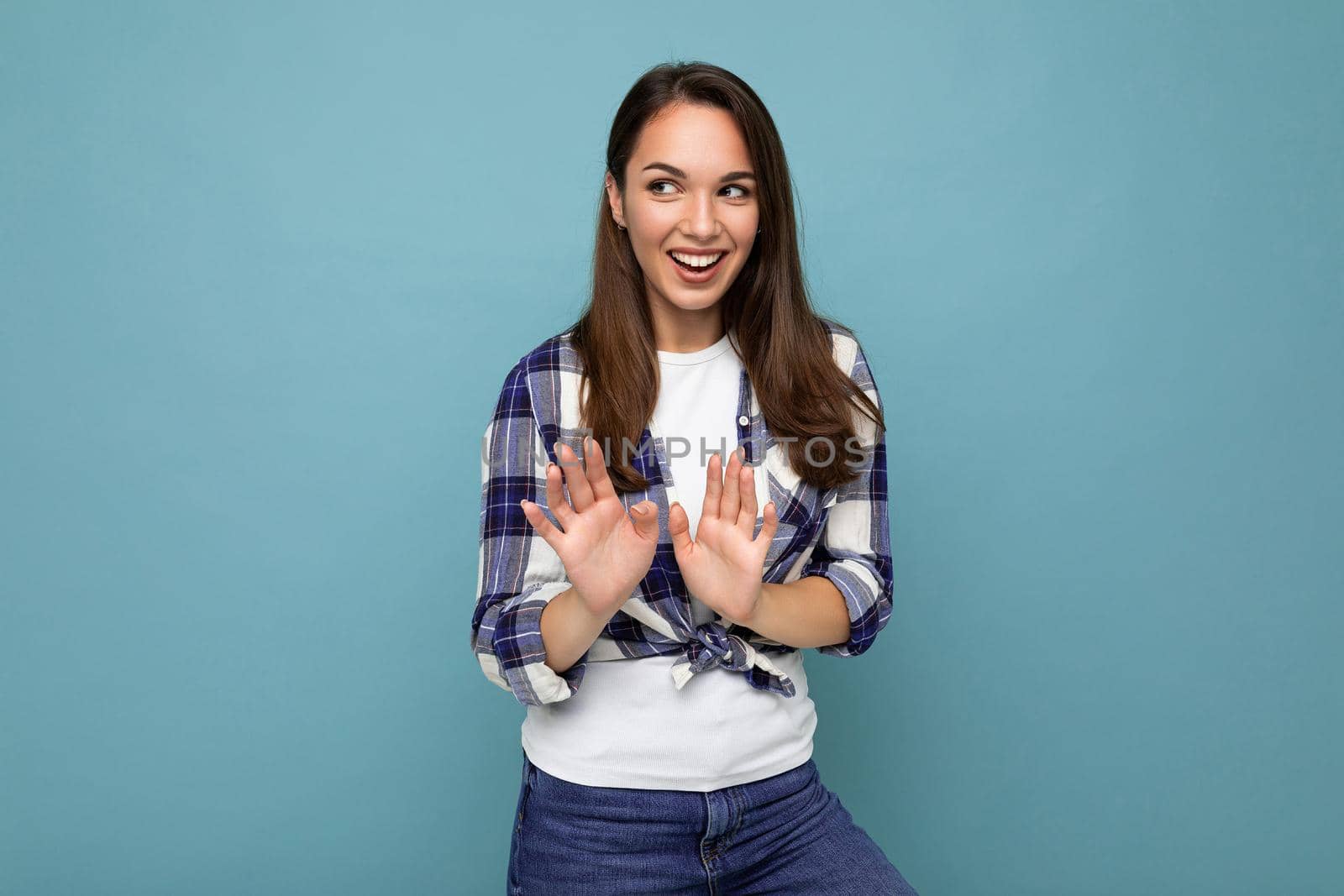 Portrait of young positive happy smiling attractive beautiful brunette woman with sincere emotions wearing hipster blue check shirt isolated over blue background with copy space and showing stop gesture.