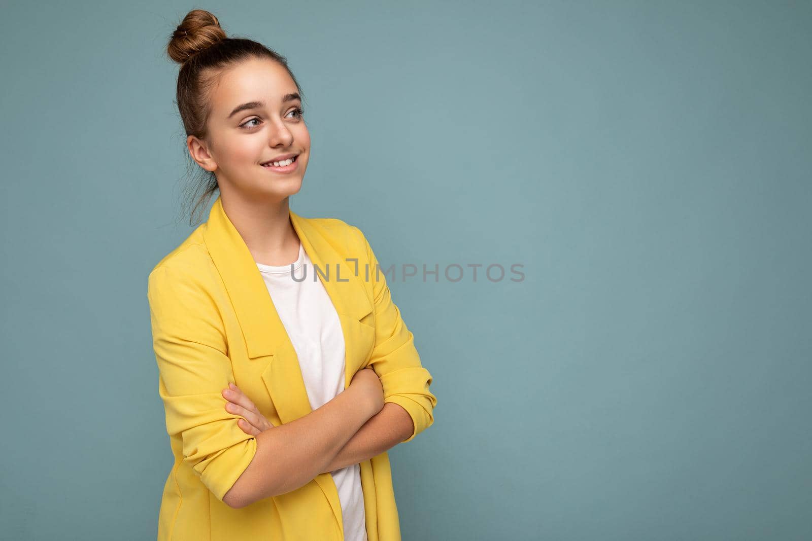 Side-profile photo shot of pretty positive happy smiling brunette little girl wearing trendy yellow jacket and white t-shirt standing isolated over blue background wall looking to the side by TRMK