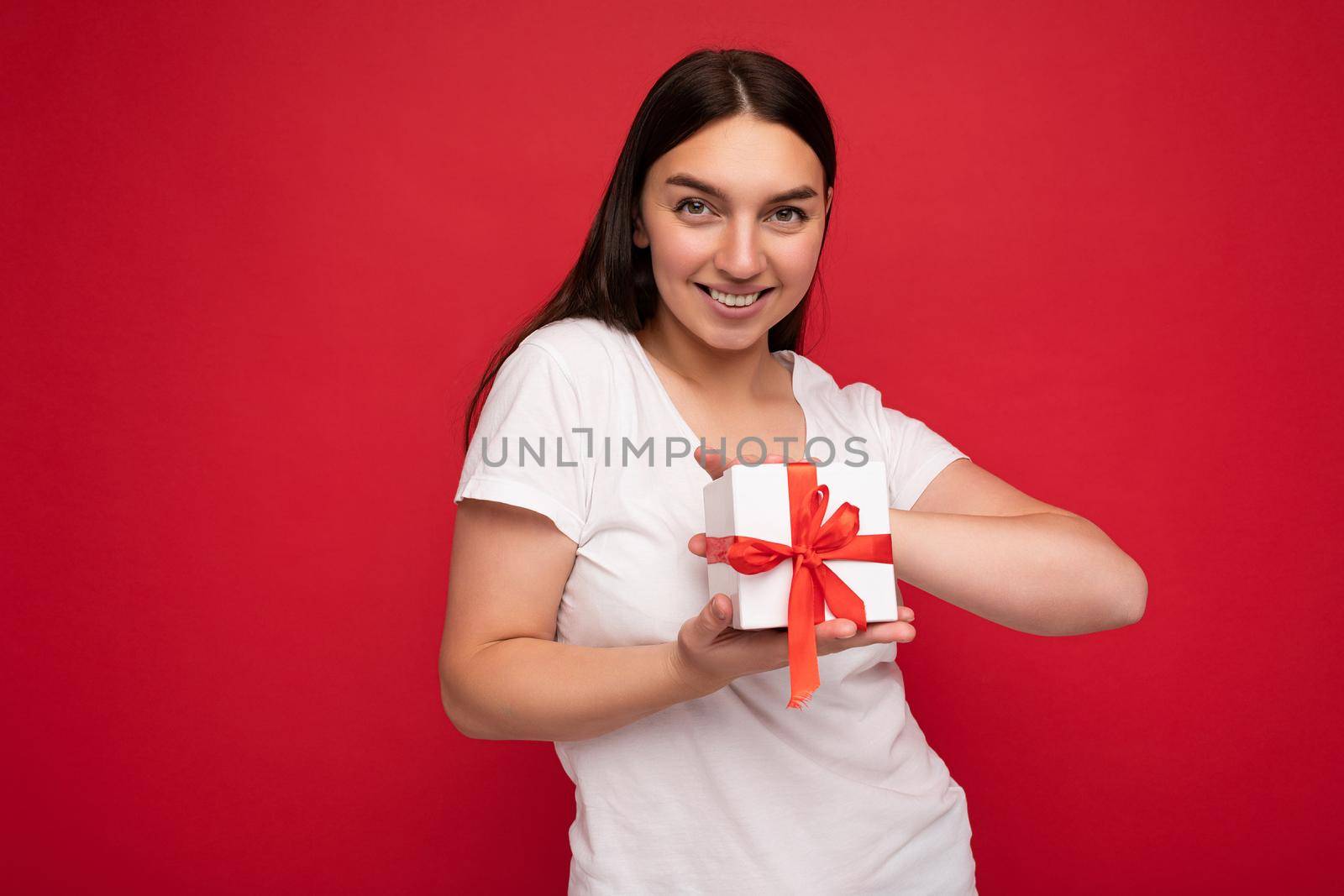 Shot of attractive positive smiling young brunette woman isolated over colourful background wall wearing everyday trendy outfit holding gift box and looking at camera.