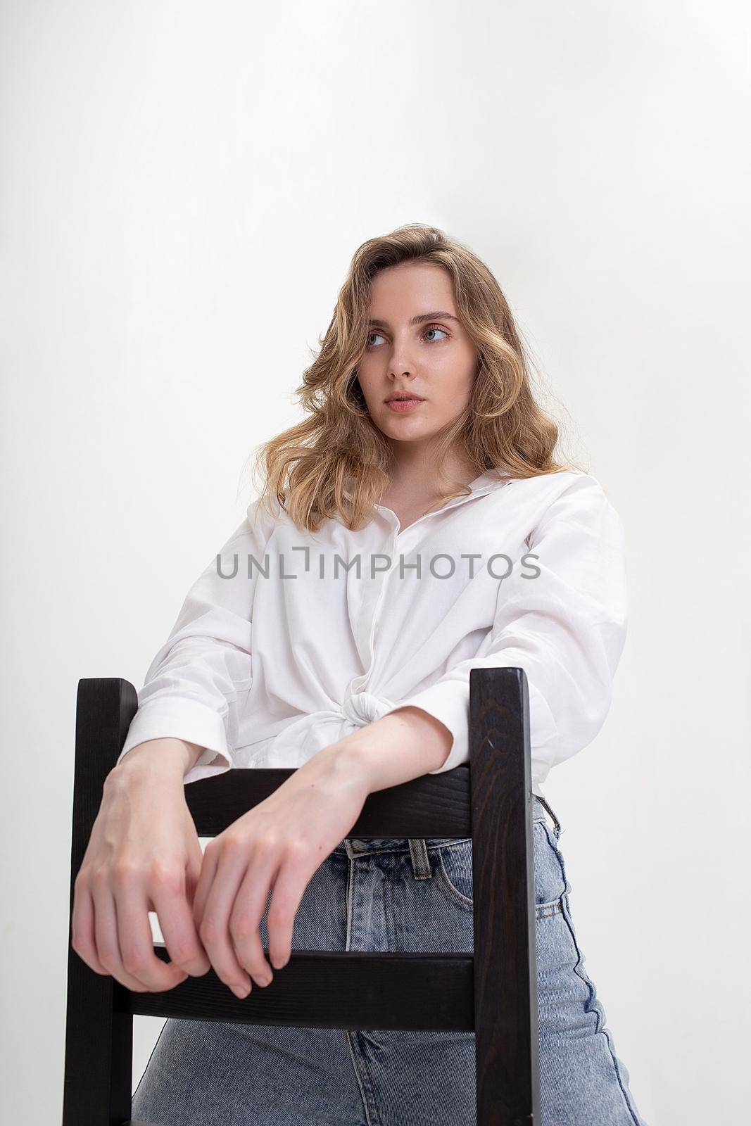 portrait of young pensive caucasian woman posing in shirt and blue jeans, sitting on black chair in white studio. model tests of pretty girl in basic clothes on cyclorama. attractive female poses