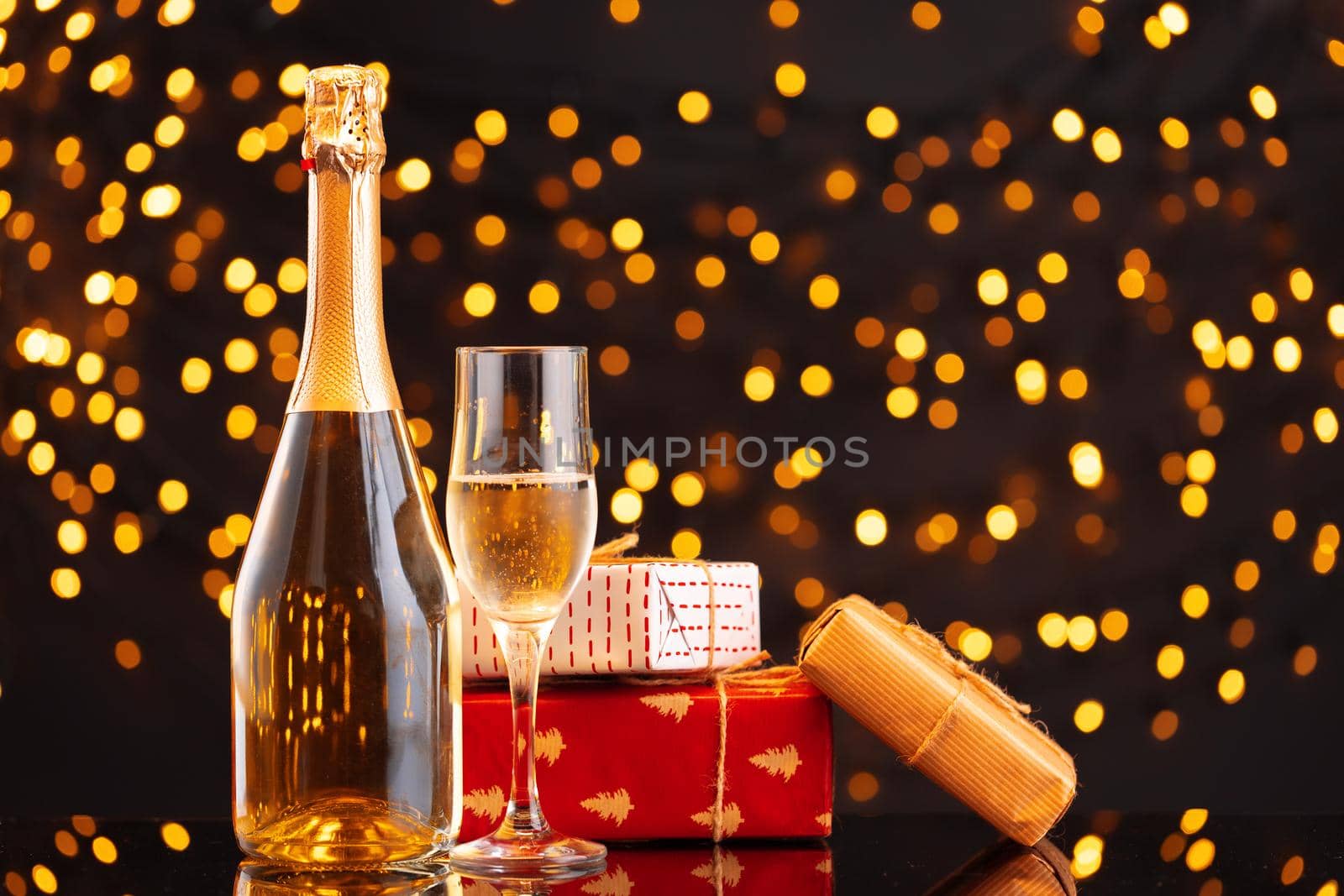 Champagne bottle and wrapped gift on blurred background of Christmas lights front view