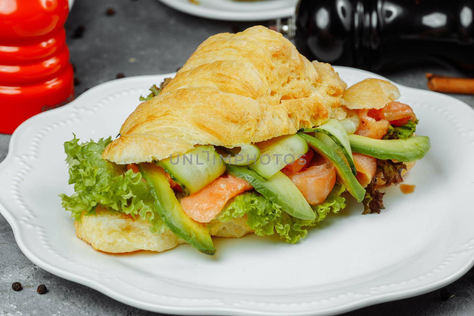 Croissant sandwich with red fish, avocado, fresh vegetables and arugula on black shale board over black stone background. Healthy food concept by UcheaD