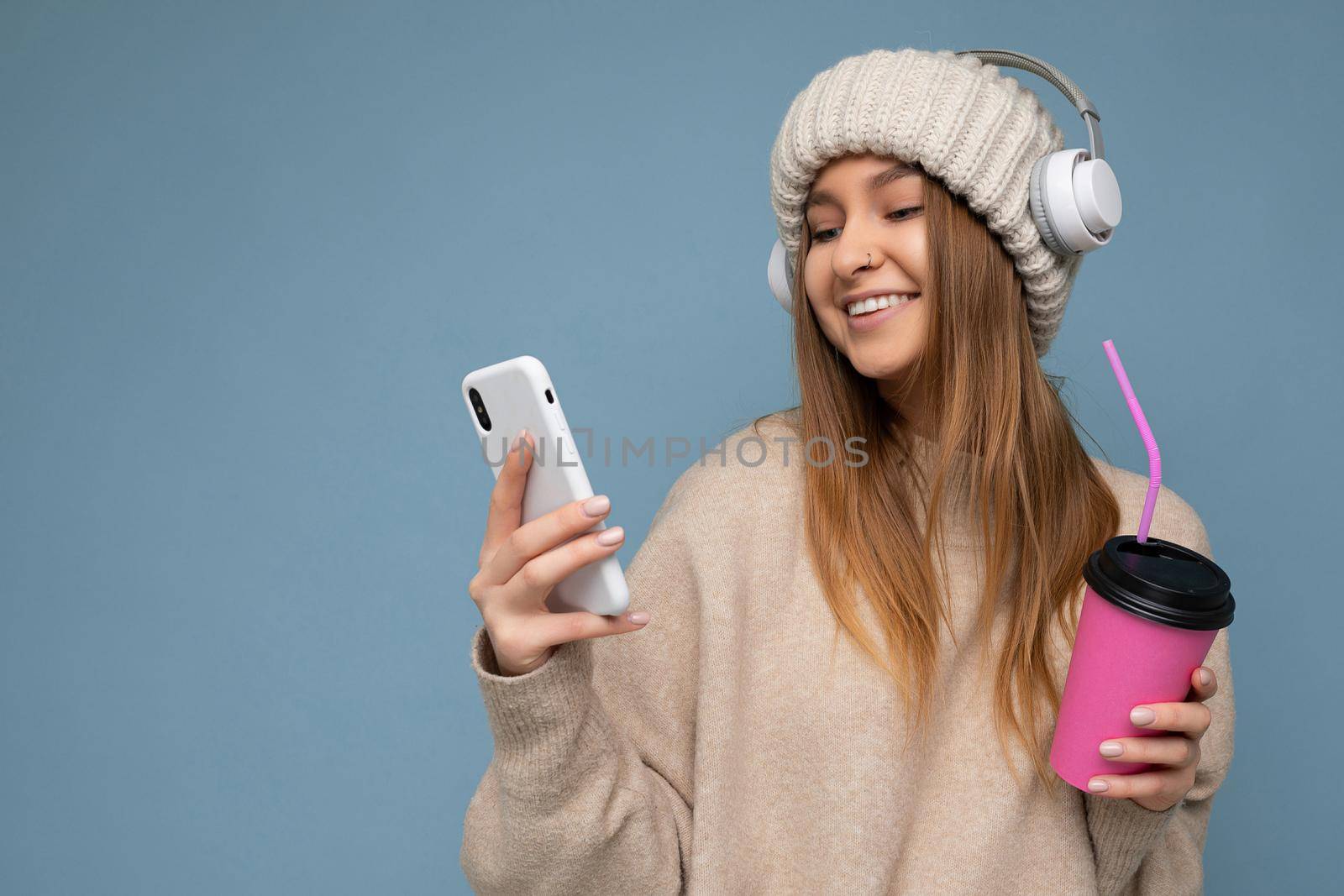 Attractive happy smiling young blonde woman wearing beige sweater and beige hat white headphones isolated over blue background holding in hand and using mobile phone reading news drinking beverage and listening to music looking at gadjet display by TRMK