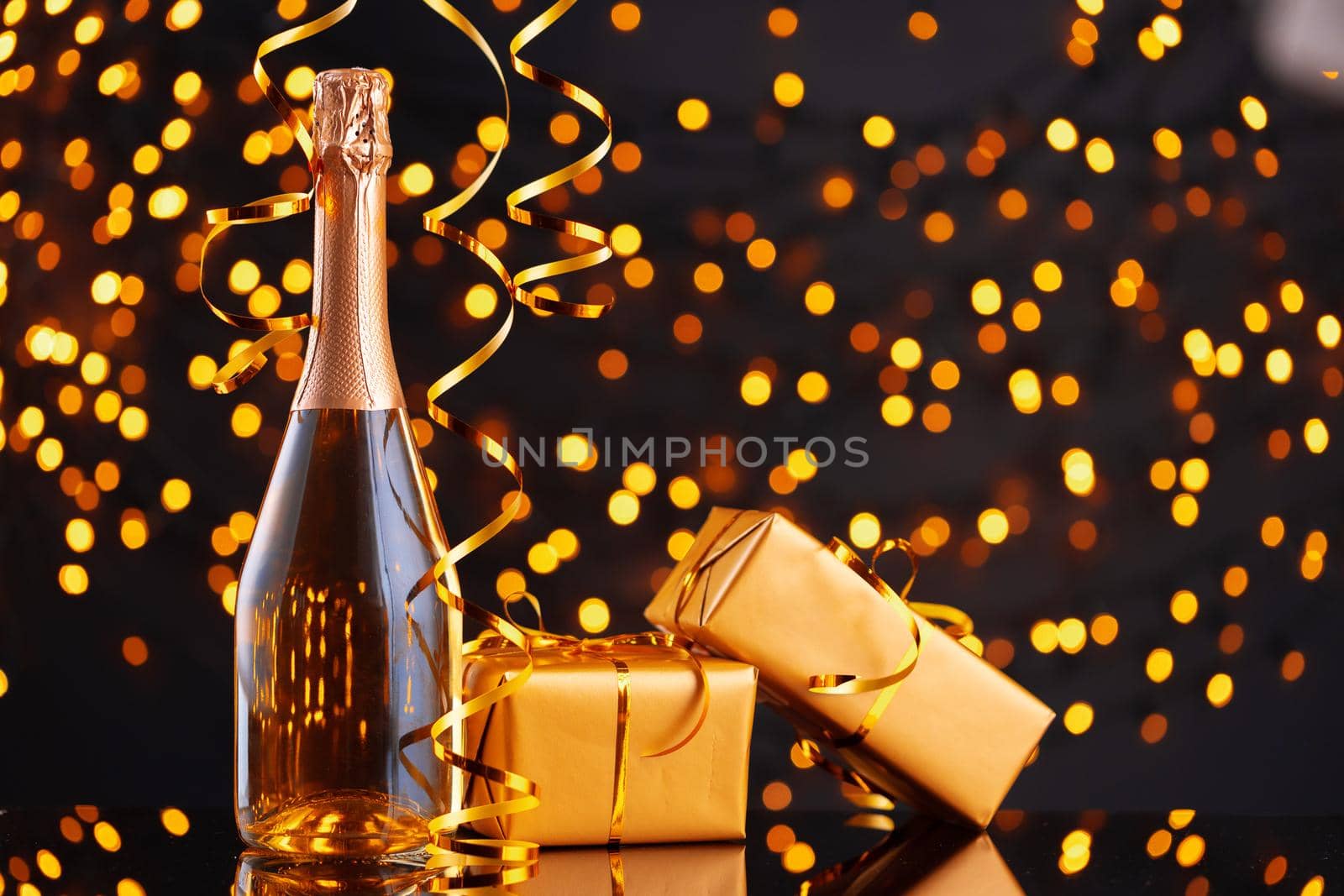 Champagne bottle and wrapped gift on blurred background of Christmas lights front view