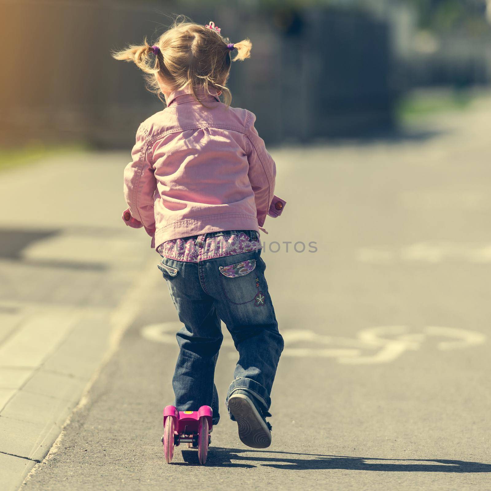 Two years old girl riding her scooter on the street. back view