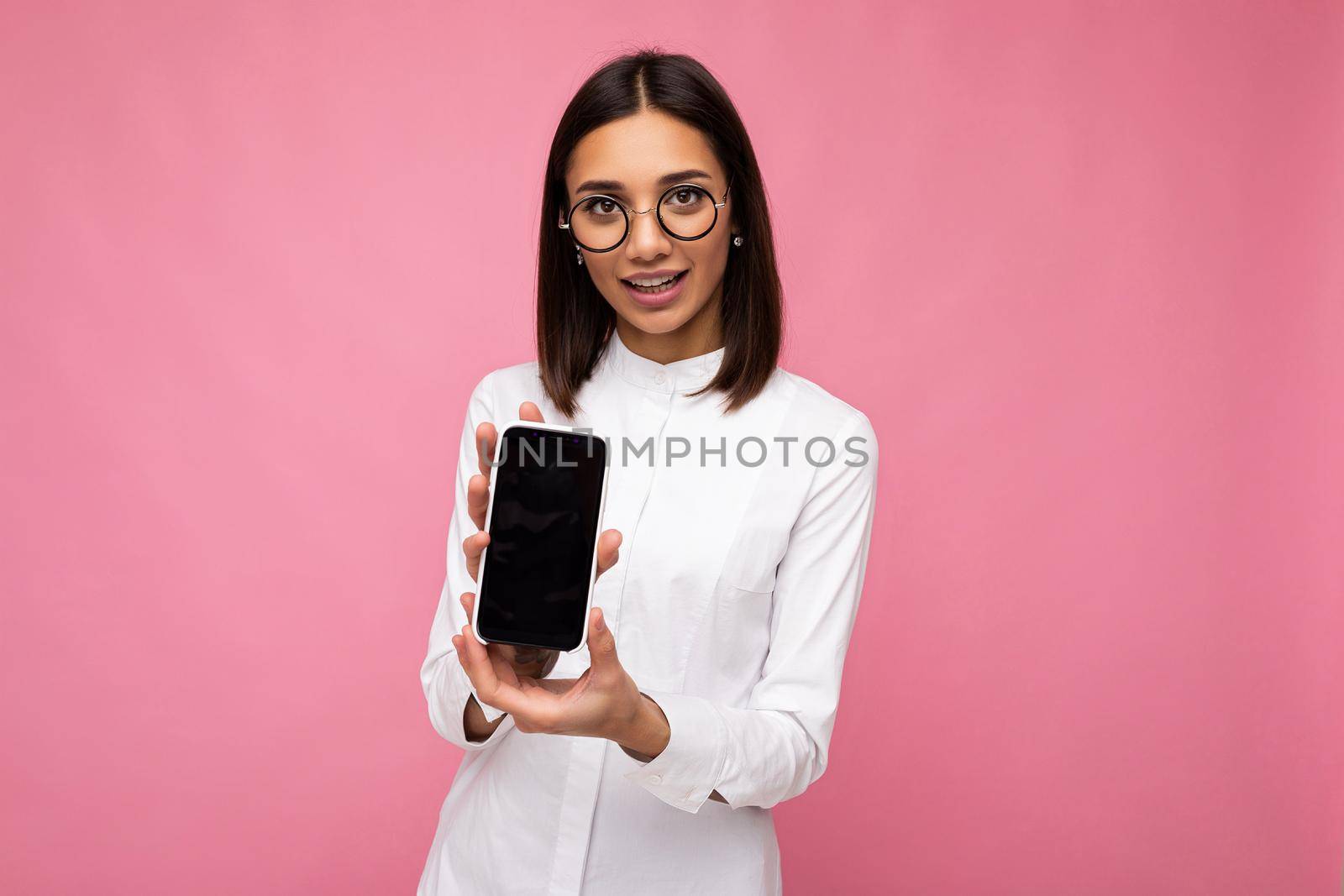 charming smiling young brunette woman wearing white blouse and optical glasses standing isolated over pink background showing mobile phone with empty screen for mockup looking at camera.