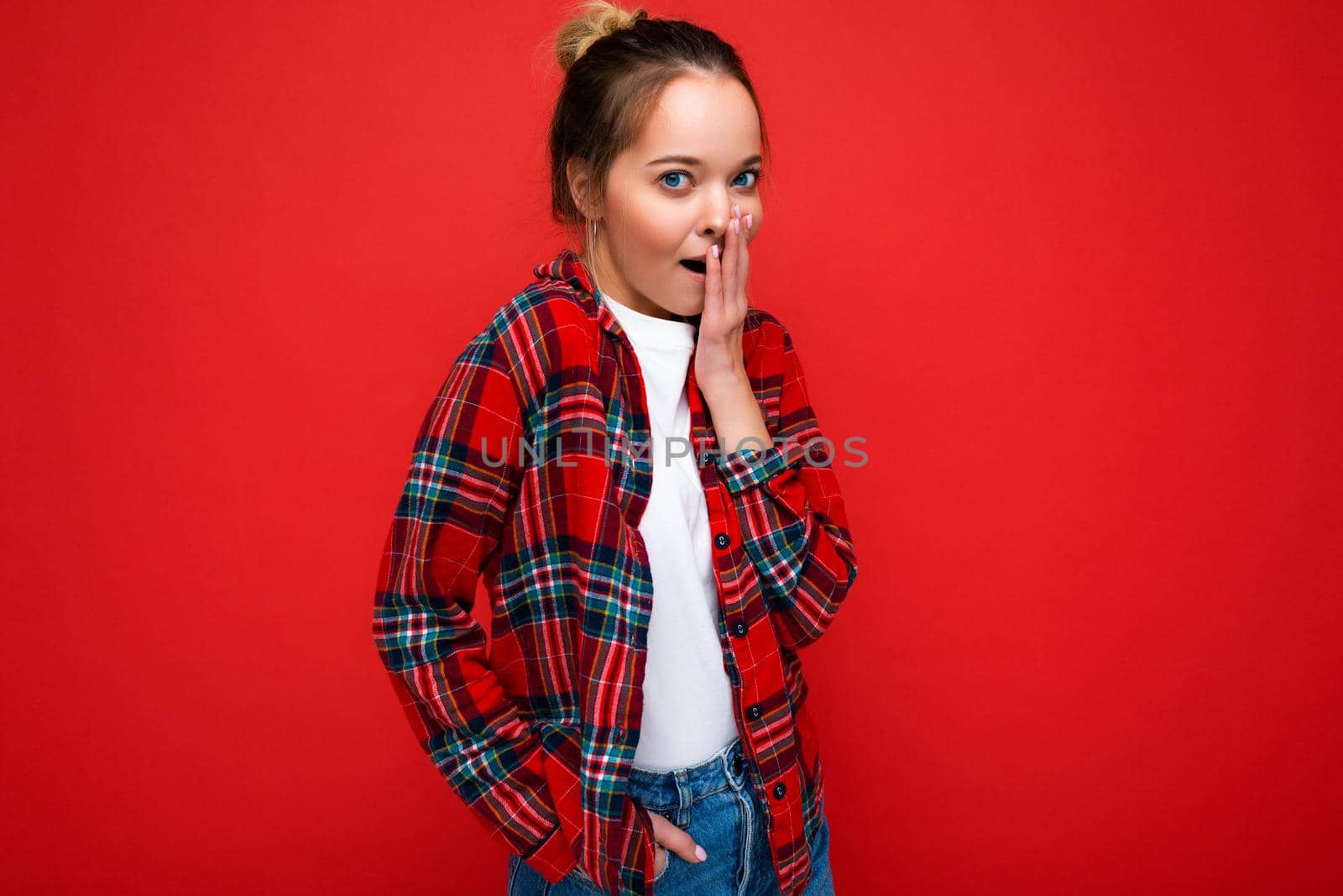 Attractive amazed young blonde woman standing isolated over red wall wearing casual red shirt and white t-shirt looking at camera. Copy space