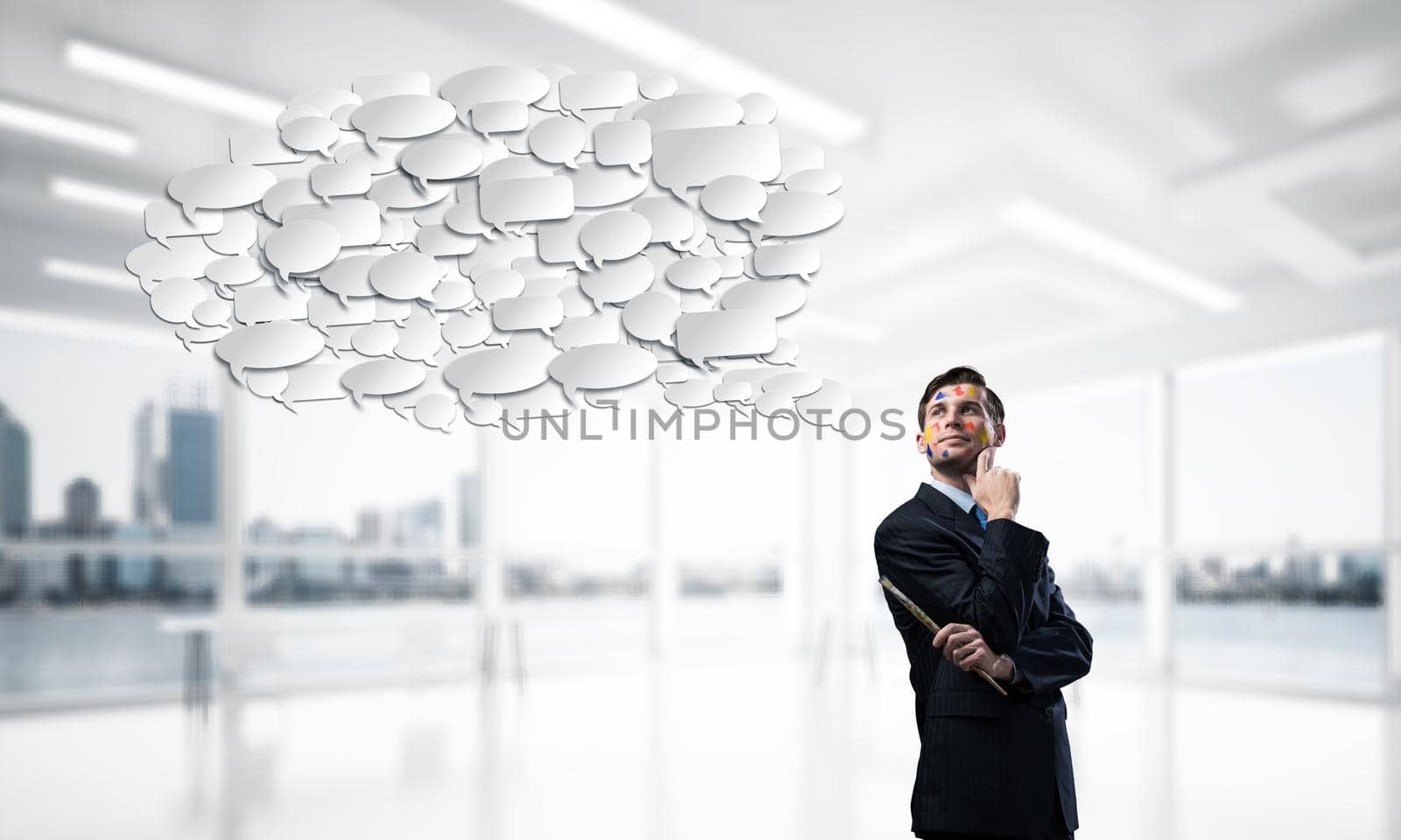 Portrait of young thoughtful man holding paintbrush in his hand and looking away while standing with chatting cloud inside modern office view on background.