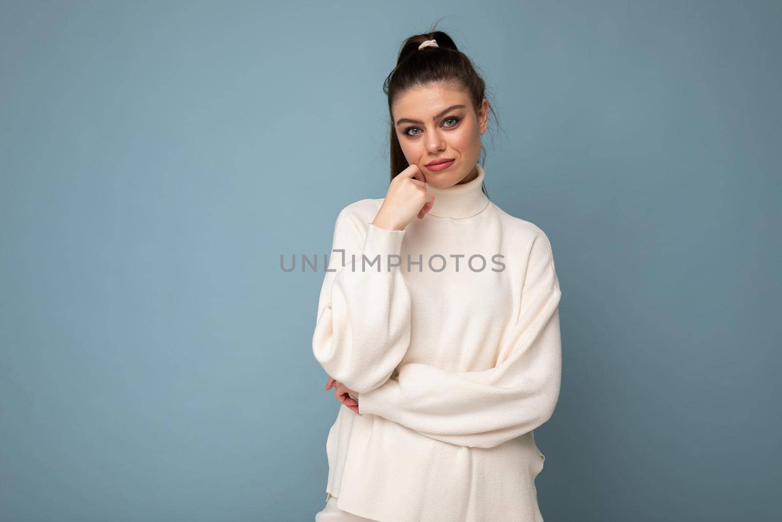 Young sad brunette woman with wearing white casual sweater isolated over blue background looking confident at the camera with crossed arms and hand raised on chin.