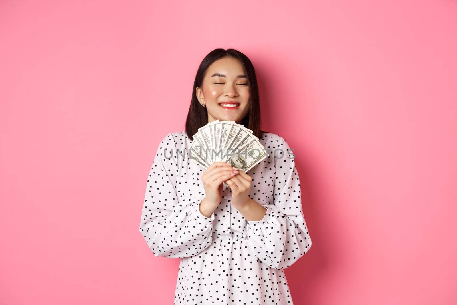 Shopping concept. Happy and satisfied asian woman winning prize money, showing dollars and rejoicing, standing over pink background.