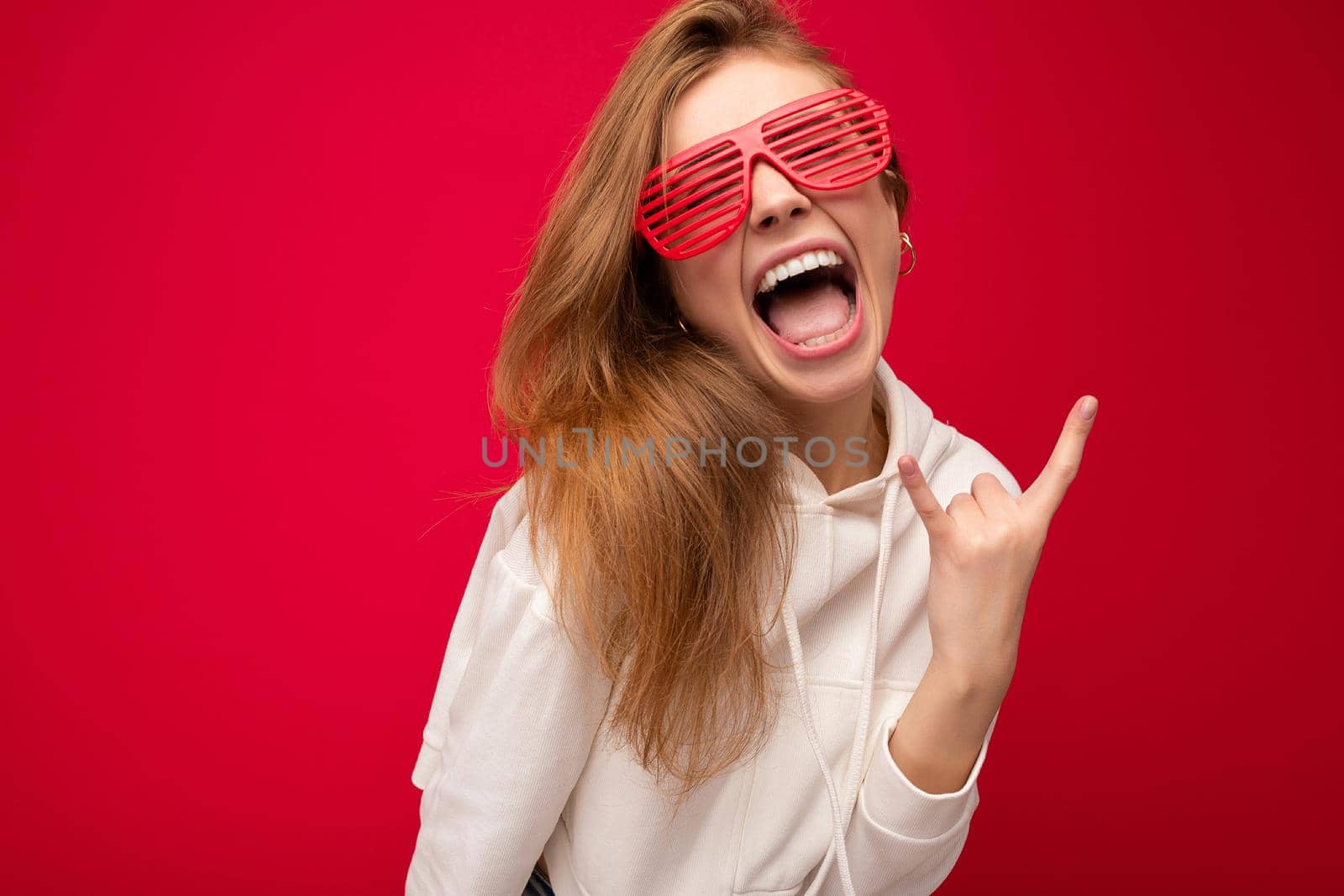 Shot of beautiful emotional young blonde woman isolated over colourful background wearing casual white hoodie and stylish sunglasses looking at camera and showing rock and roll tongue.