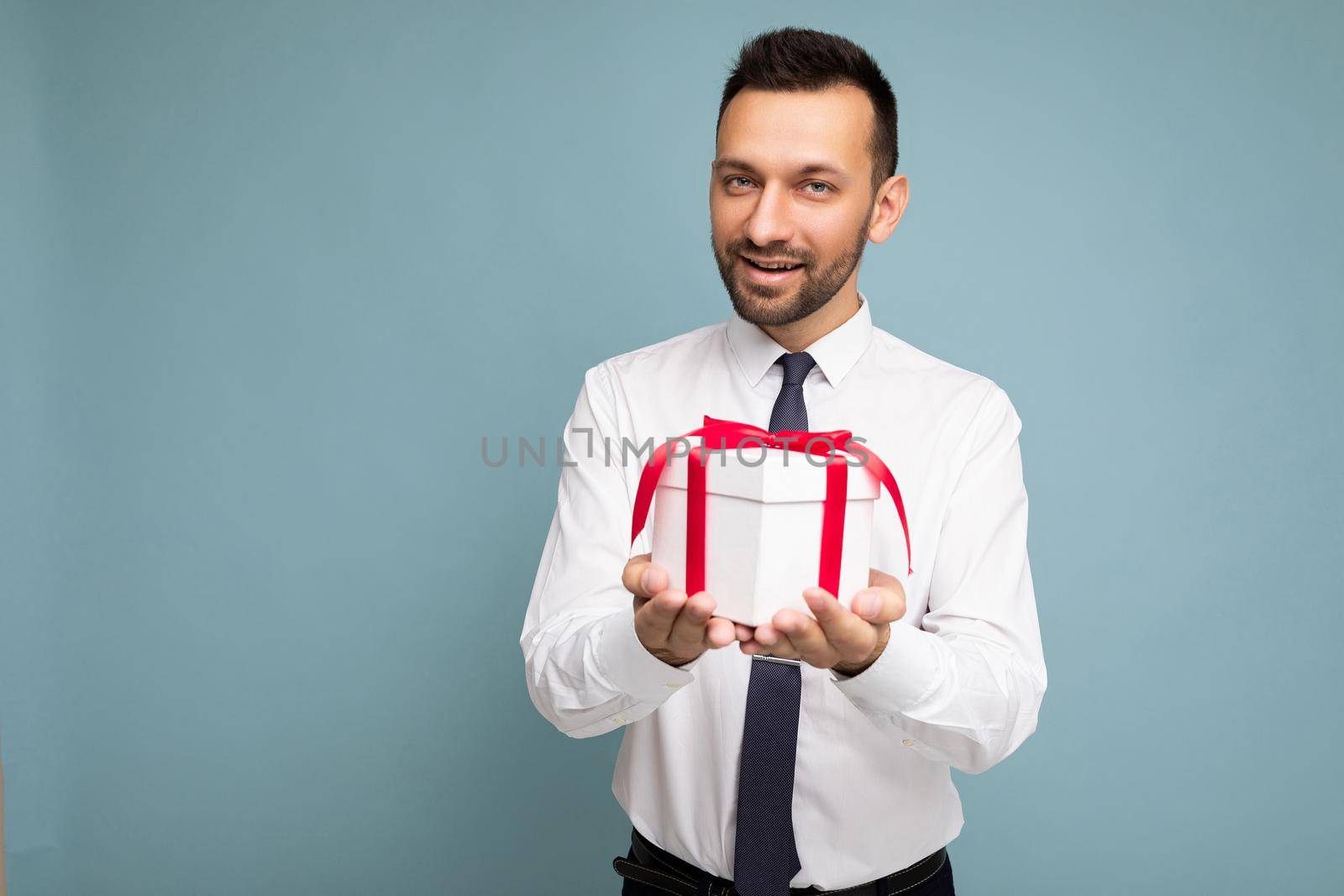 Handsome positive brunette young unshaven businessman male peson with beard isolated over blue background wall wearing white shirt and tie holding white gift box with red ribbon and looking at camera. Free space