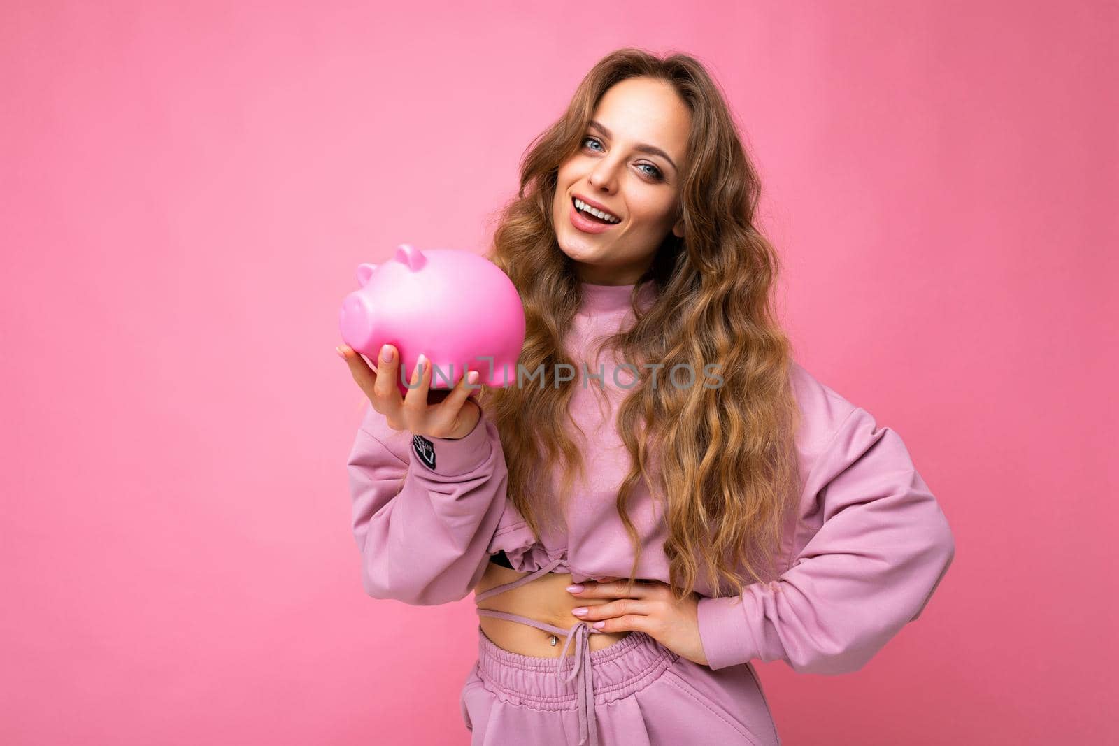 Portrait of happy positive smiling young beautiful attractive woman with wavy long blonde hair with sincere emotions wearing stylish pink hoodie isolated over pink background with copy space and holding pink piggy box. Money box concept by TRMK