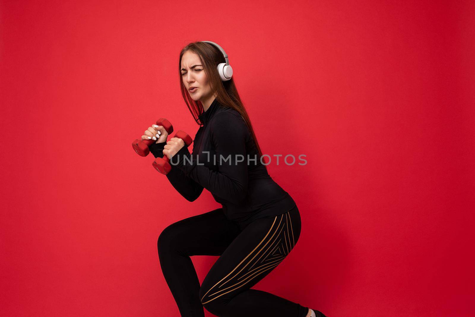 Beautiful happy smiling young brunet woman wearing black sport clothes isolated on red background wall doing fitness using dumbbells wearing white bluetooth headsets listening to music looking at camera.