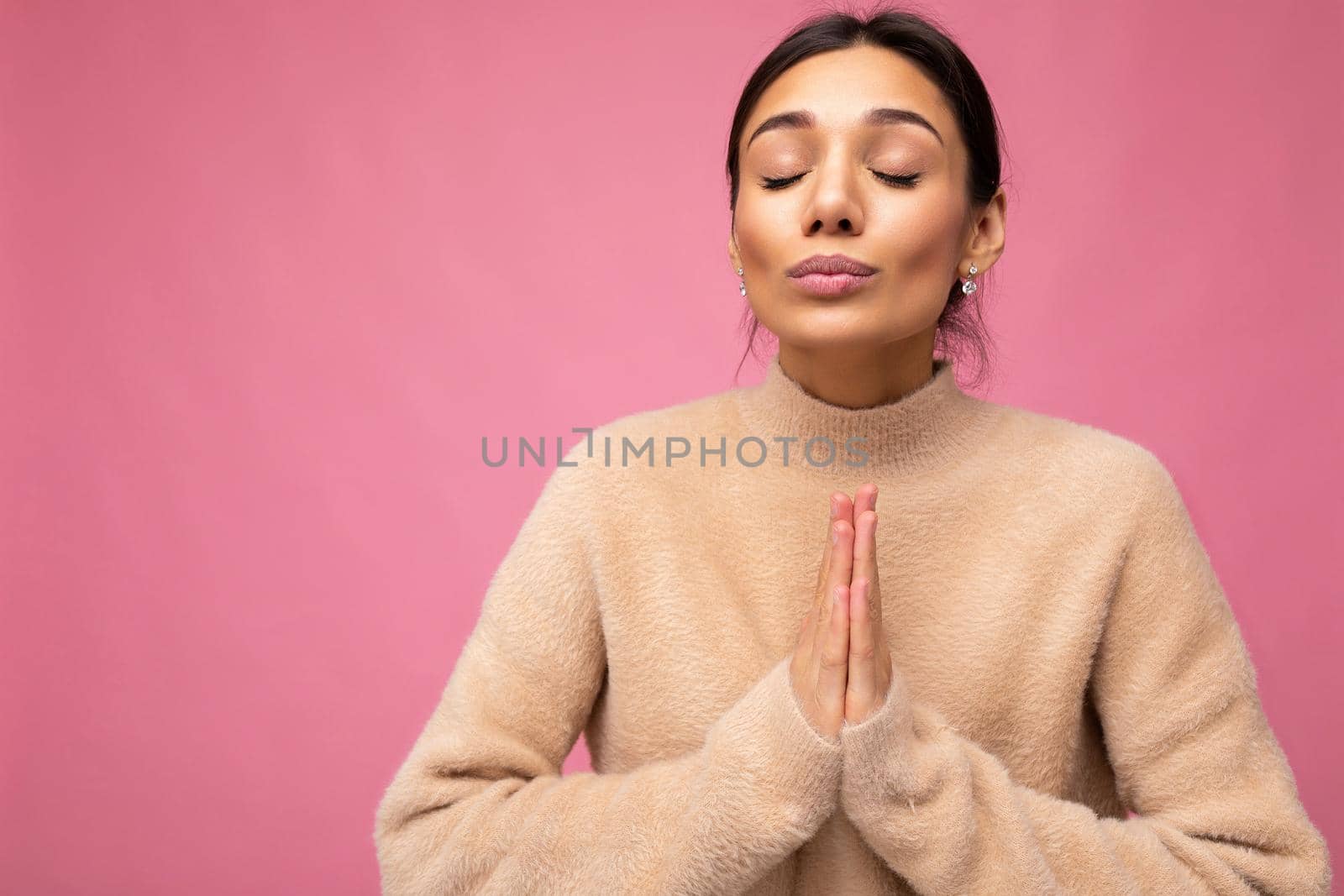 Portrait of young positive happy beautiful brunette woman with sincere emotions wearing casual beige jersey isolated over pink background with free space and holding hands in praying gesture by TRMK