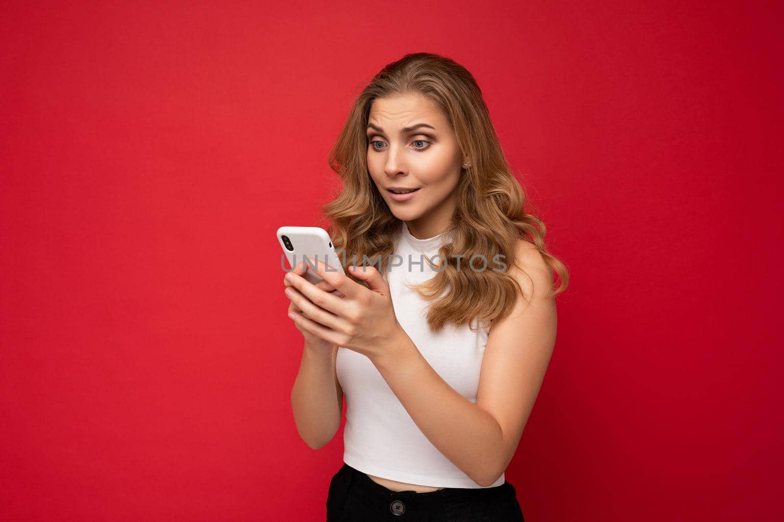 Amazed shocked beautiful young blonde woman wearing white t-shirt isolated over red background using smartphone and texting message via mobile phone looking at gadjet screen by TRMK