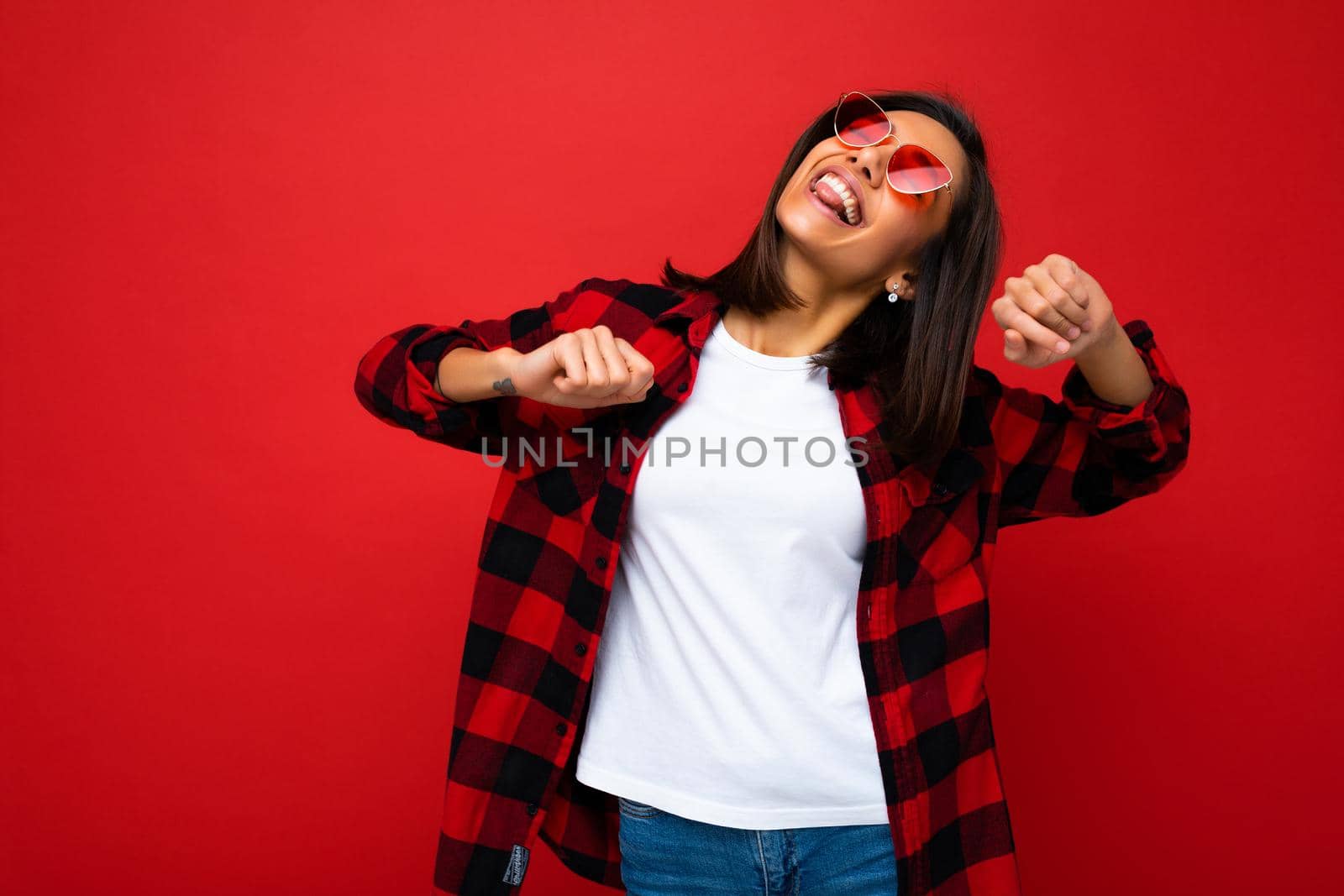 Portrait of young happy positive beautiful brunette woman with sincere emotions wearing white t-shirt, stylish red check shirt and red sunglasses isolated on red background with copy space and having fun.