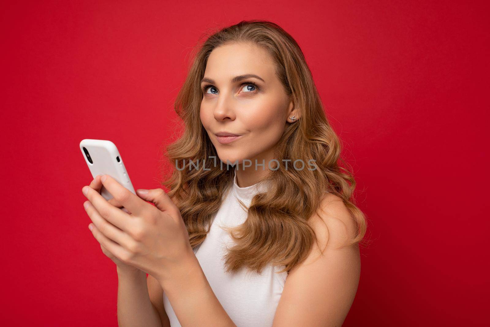 Closeup of thoughtful happy pretty young blonde woman wearing white t-shirt isolated over red background using smartphone and chatting via mobile phone looking to the side and dreaming.