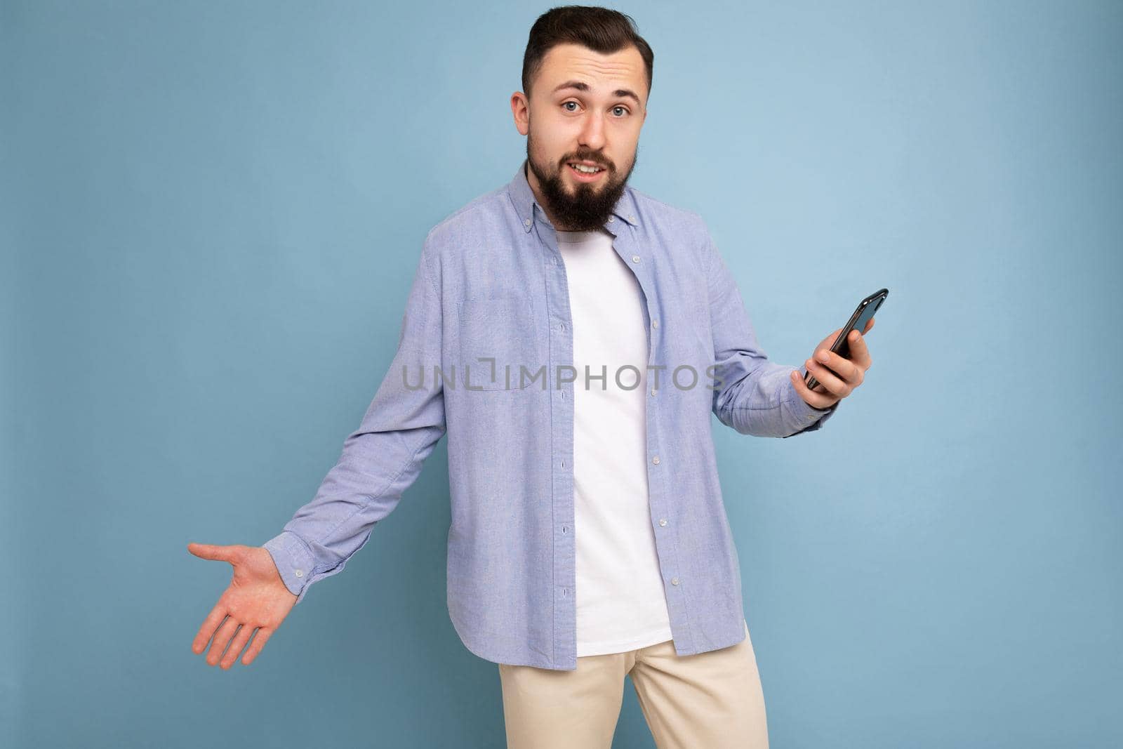 Upset handsome young brunette unshaven man with beard wearing stylish white t-shirt and blue shirt isolated over blue background with empty space holding in hand and using phone messaging sms looking at camera and having troubles.