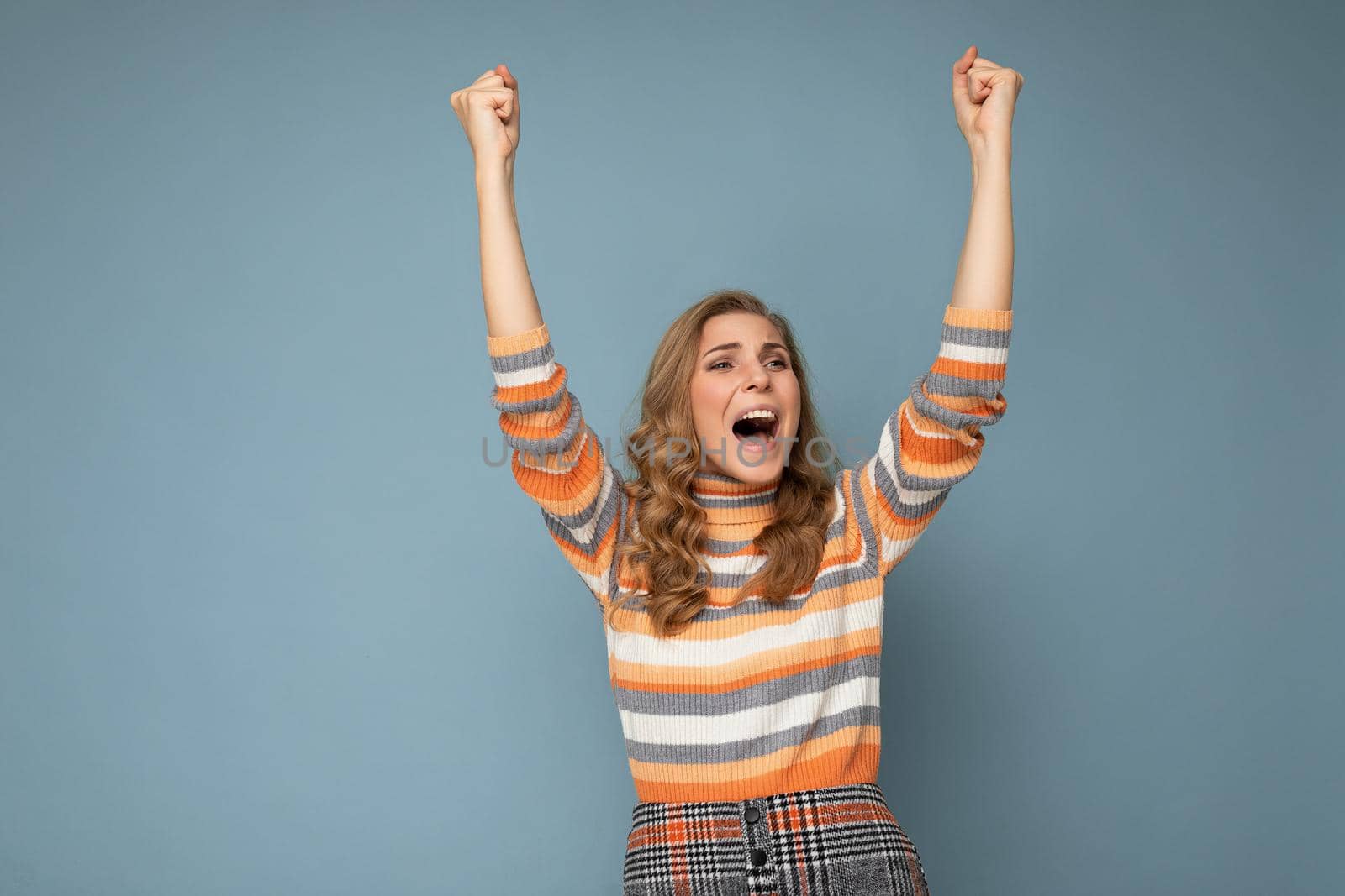 Photo portrait of young pretty beautiful happy positive funny joyful blonde woman with sincere emotions wearing striped pullover isolated on blue background with empty space and celebrating winning shouting yeah. Victory concept by TRMK