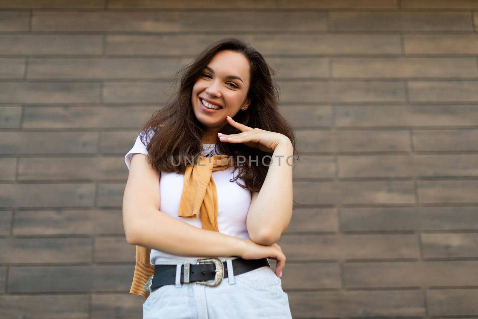 Portrait of successful smiling joyful happy young brunet woman wearing casual white t-shirt and jeans with yellow sweater poising near brown wall in the street and having fun.