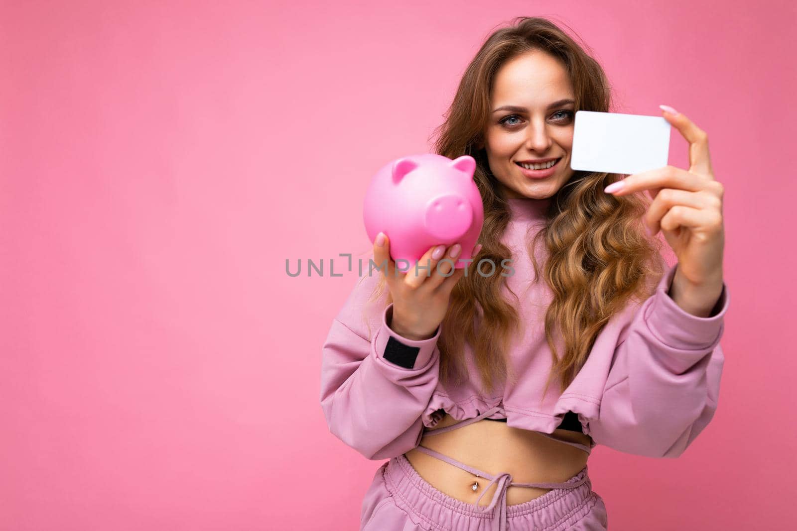 Shot of happy positive smiling young winsome attractive woman with wavy long blonde hair with sincere emotions wearing stylish pink hoodie isolated over pink background with copy space, holds pink pig penny bank for money and credit card. Savings concept by TRMK