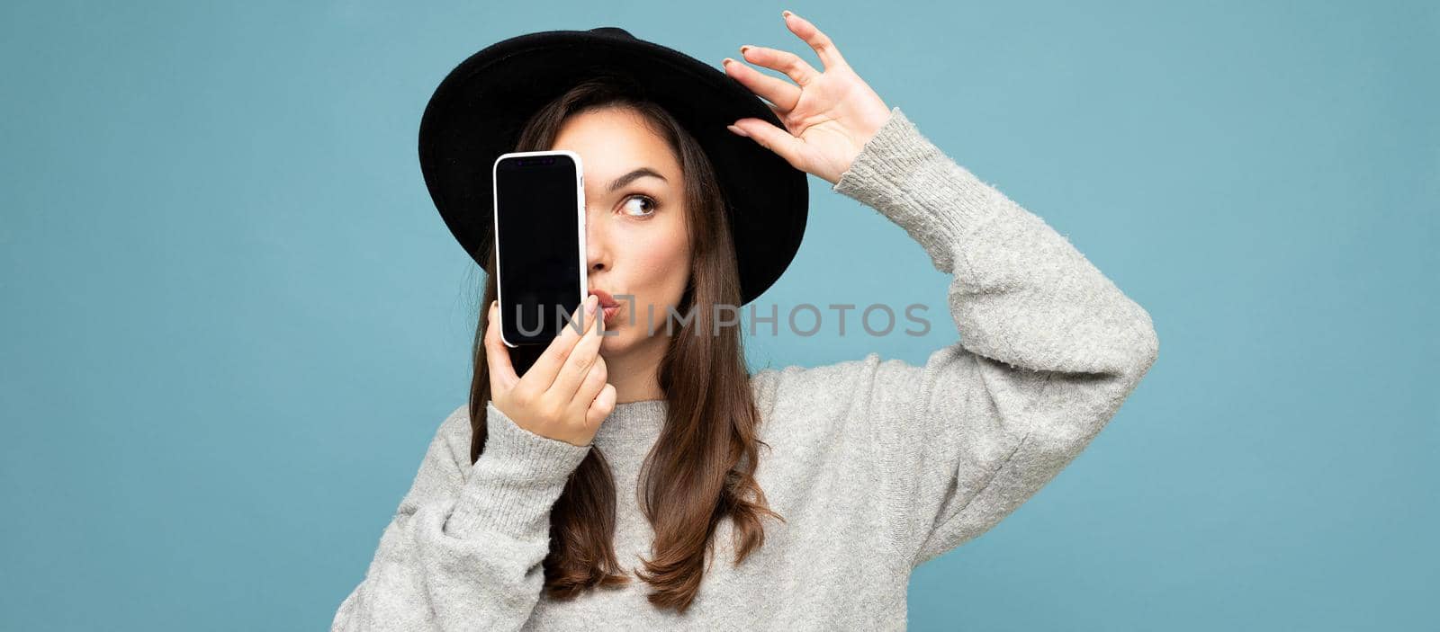 panoramic Photo of Beautiful amazed positive woman wearing black hat and grey sweater holding mobilephone showing smartphone isolated on background looking to the side.Mock up, cutout, free space. copy space