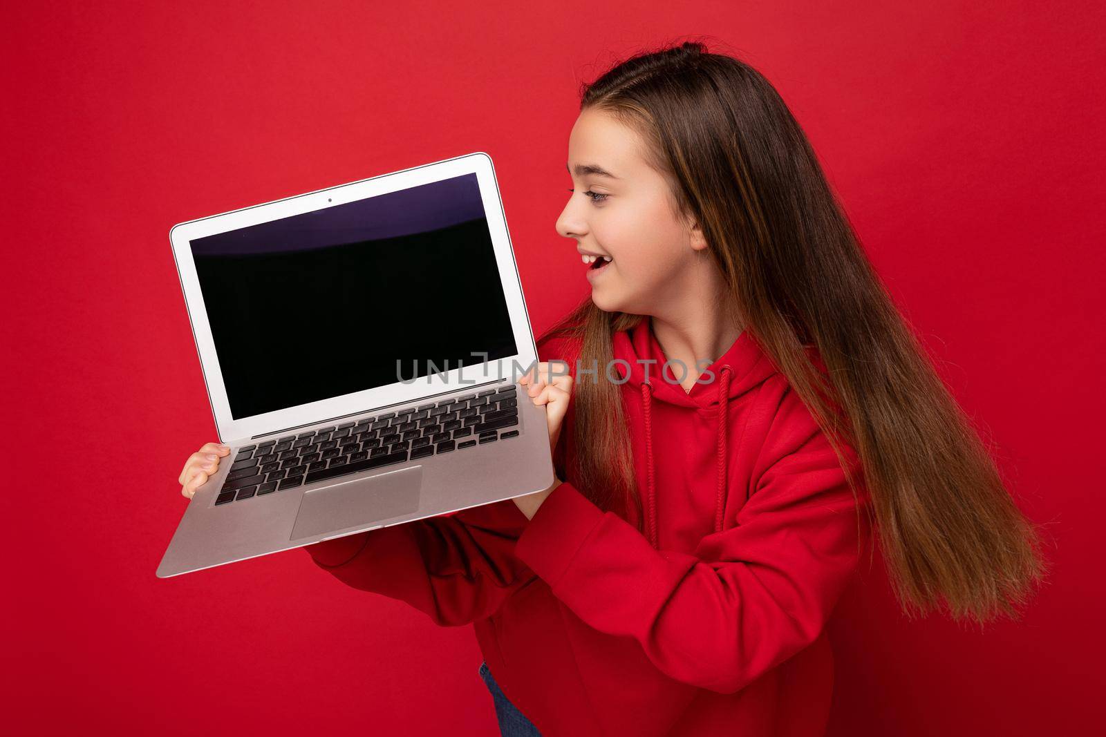 Side profile photo of beautiful happy girl with long hair wearing red hoodie holding computer laptop looking at netbook keyboard and screen monitor isolated over red wall. Cutout