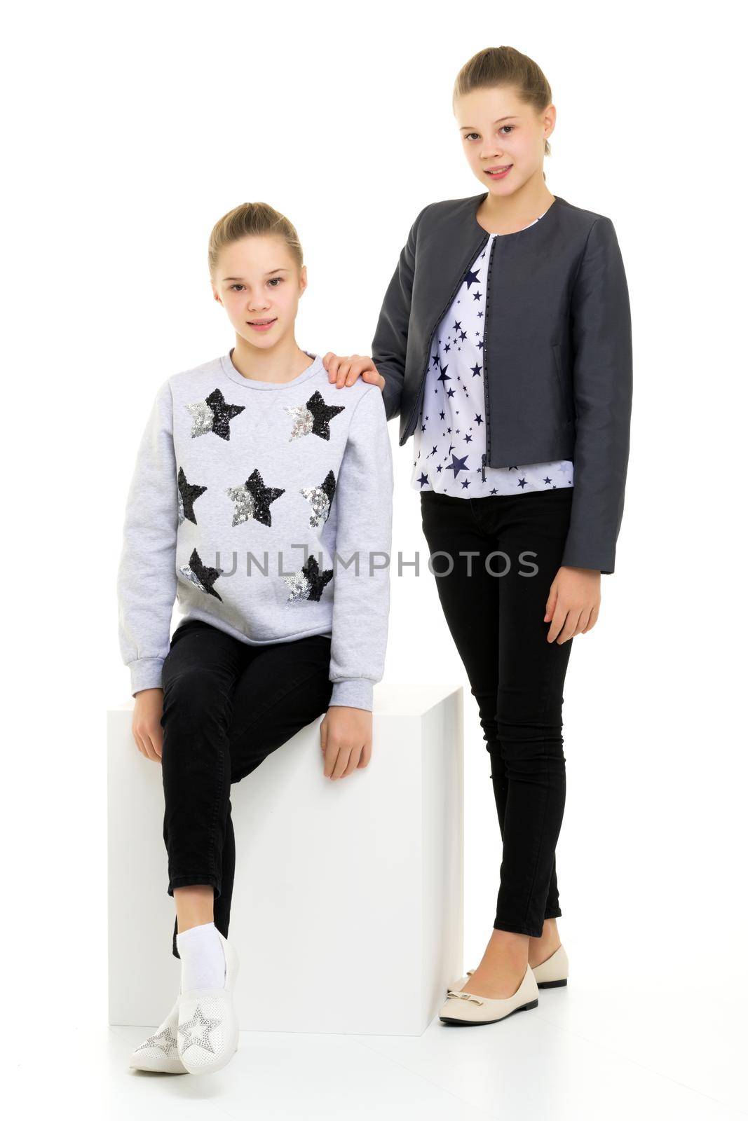 Pretty Sisters Standing Leaning on Cube with one Knee and Looking at Camera, Full Length Portrait of Two Teen Girls Wearing Trendy Stylish Clothes Posing Together in Studio Against White Background