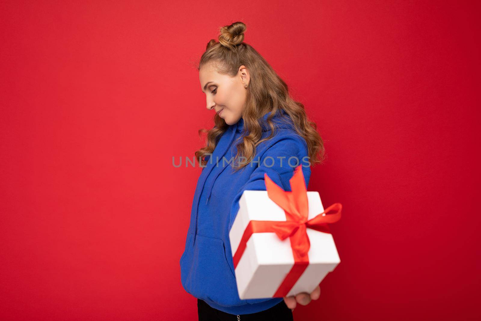 Shot of attractive happy young blonde woman isolated over red background wall wearing blue trendy hoodie holding white gift box with red ribbon and looking to the side by TRMK
