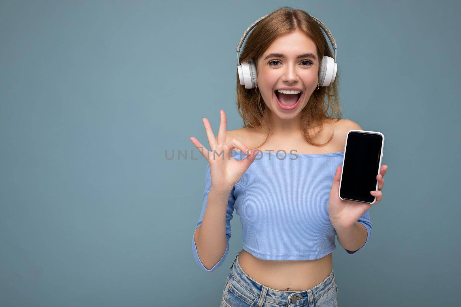 Attractive positive smiling young woman wearing stylish casual outfit isolated on colourful background wall holding and showing mobile phone with empty screen for cutout wearing white bluetooth headphones and having fun looking at camera and showing ok gesture.