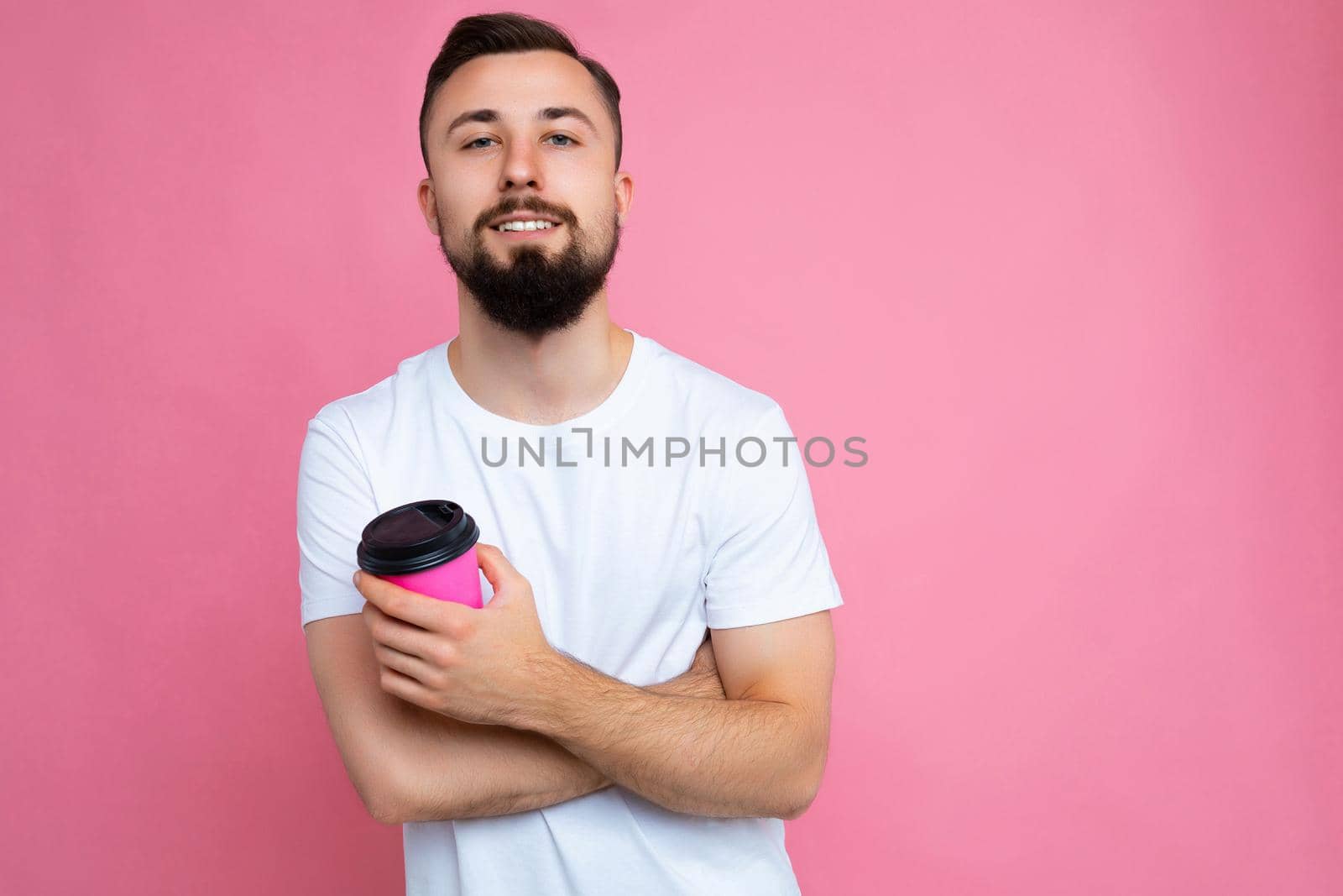 Self-confident handsome happy young brunette unshaven man with beard wearing white t-shirt isolated over pink backgroung wall holding paper coffee cup drinking and looking at camera by TRMK