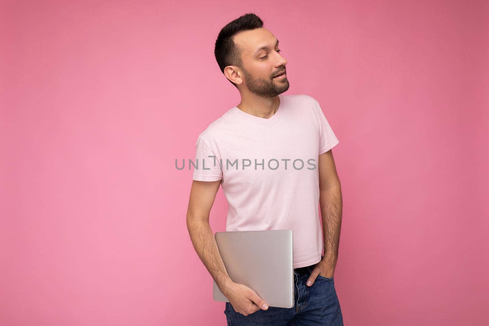 Handsome happy young unshaven man holding laptop computer looking to side in t-shirt on isolated pink background.