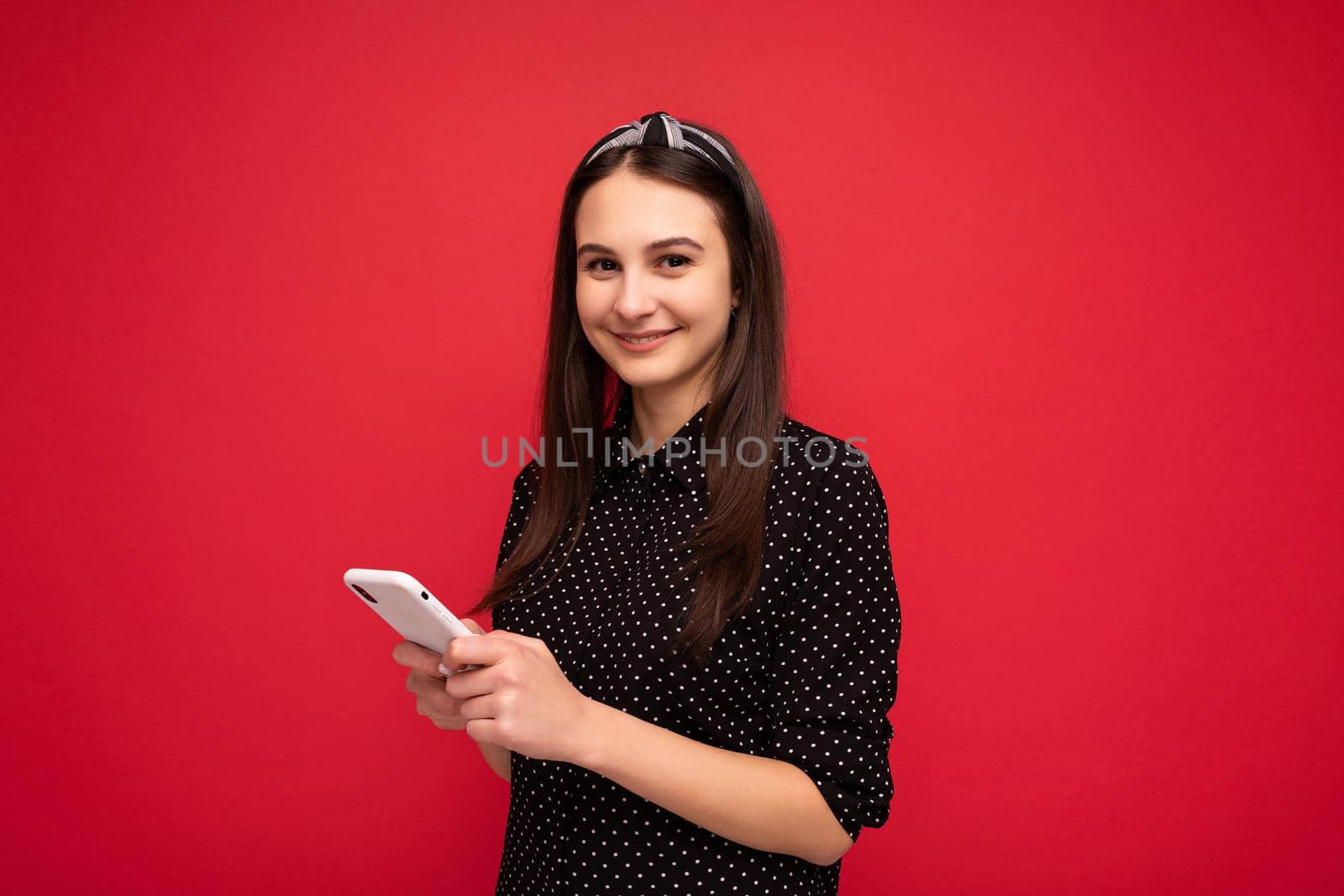 Photo shot of attractive positive good looking young lady wearing casual stylish outfit poising isolated on background with empty space holding in hand and using mobile phone messaging sms looking at camera.