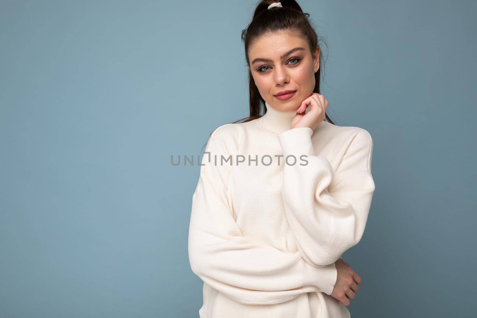 Young brunette woman with wearing white casual sweater isolated over blue background looking confident at the camera with crossed arms and hand raised on chin. Thinking positive by TRMK