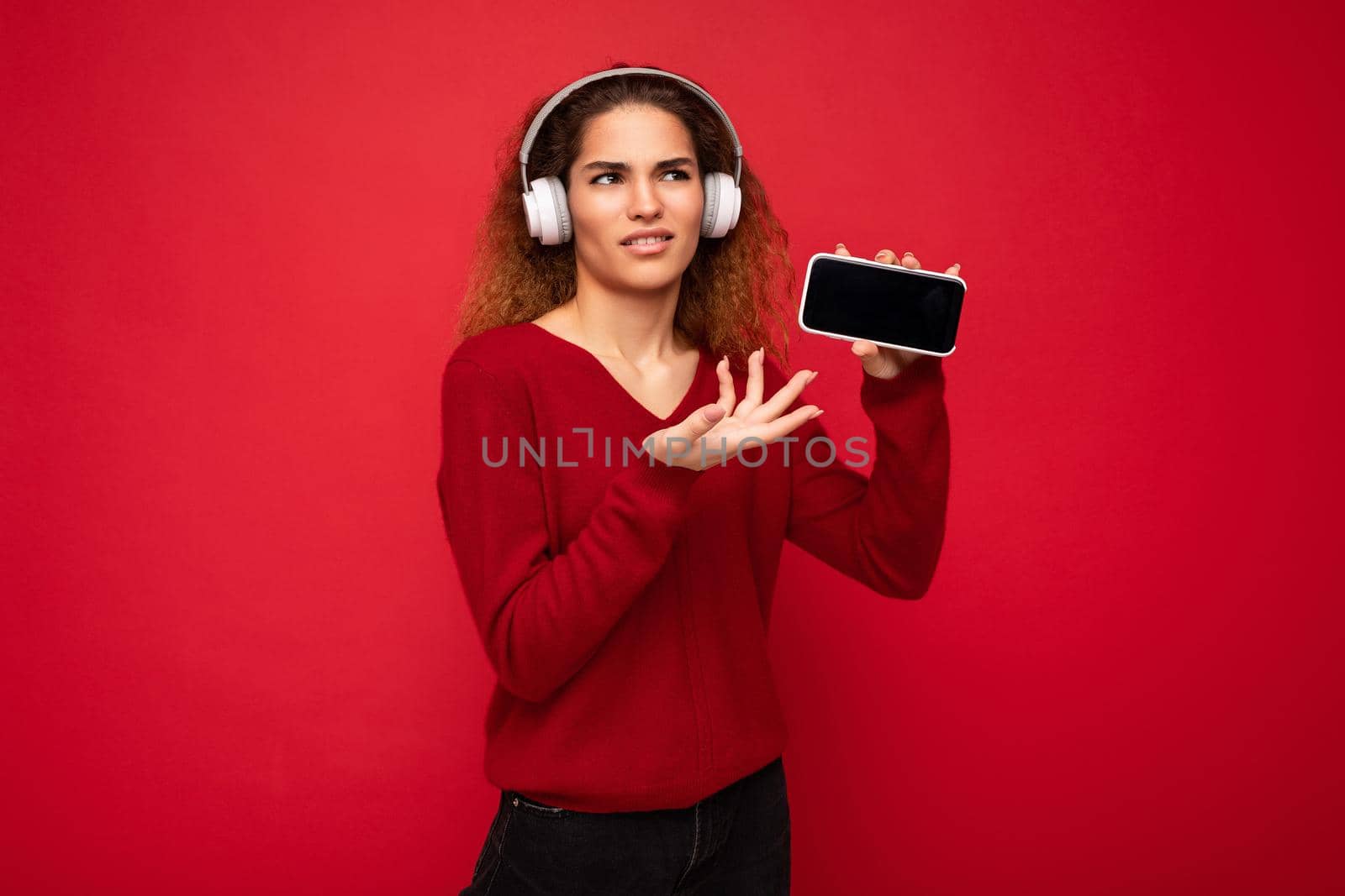 Attractive dissatisfied young woman wearing stylish casual outfit isolated on colourful background wall holding and showing mobile phone with empty screen for cutout wearing white bluetooth headphones.