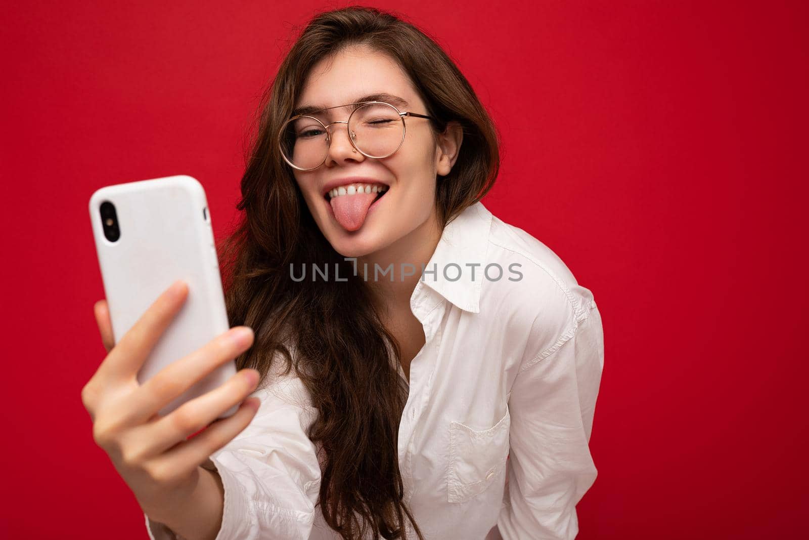 Closeup photo of sexy beautiful positive young brunette woman wearing white shirt and optical glasses isolated over red background holding in hand and using mobile phone taking selfiie looking at gadjet screen and showing tongue.
