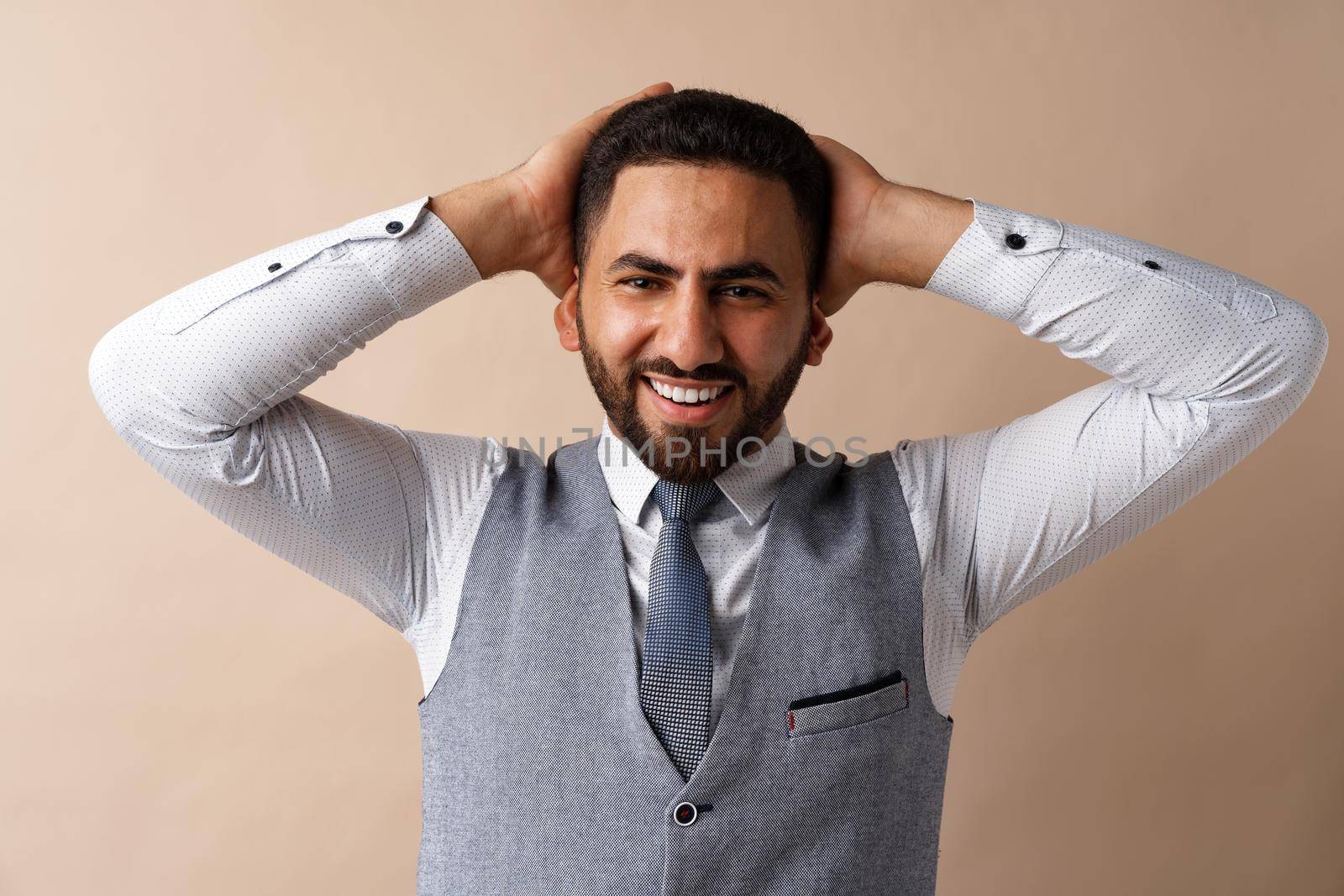 Smiling young man with hands behind head against beige background, close up