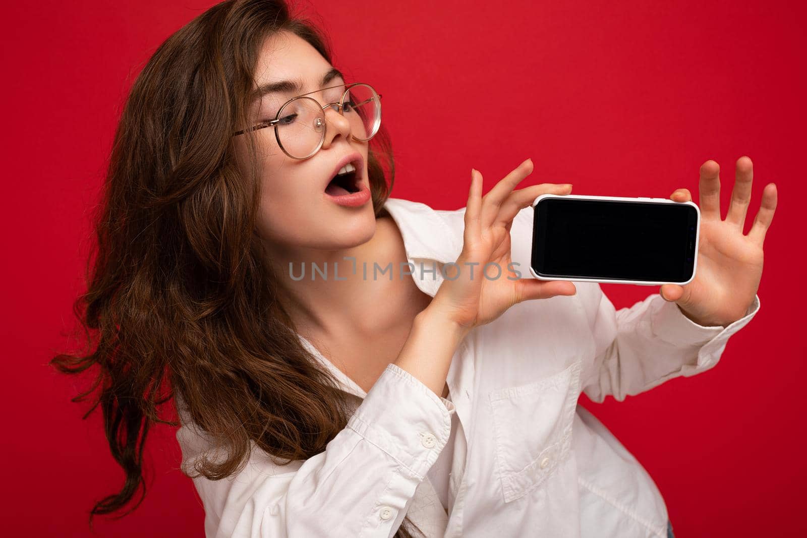 photo of sexy shocked beautiful young brunette woman wearing white shirt and optical glasses isolated over red background holding in hand and showing mobile phone with empty screen for cutout looking at gadjet screen by TRMK