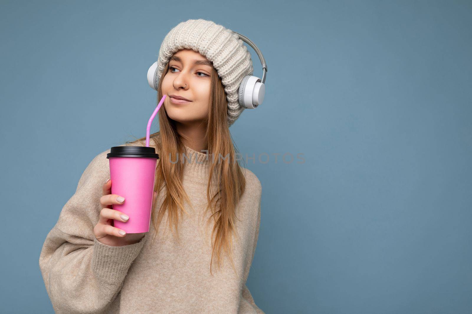 Photo shot of Beautiful young happy blonde woman wearing knitted hat beige sweater and white wireless headsets listening to music isolated over blue background holding paper tes cup for mockup drinking and looking to the side. copy space